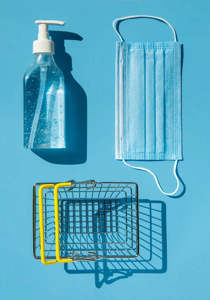 Personal protection kit for shopping during the coronavirus pandemic: face mask, gel sanitizer and shopping basket. Blue background, contrasting shadows. The view from the top.