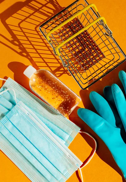 Personal protection kit for shopping during the coronavirus pandemic: individual face masks, gel sanitizer, hand gloves and shopping basket. Orange background, contrasting shadows. The view from the top.