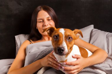 Genç güzel esmer kadın yatakta köpeğiyle oynuyor.