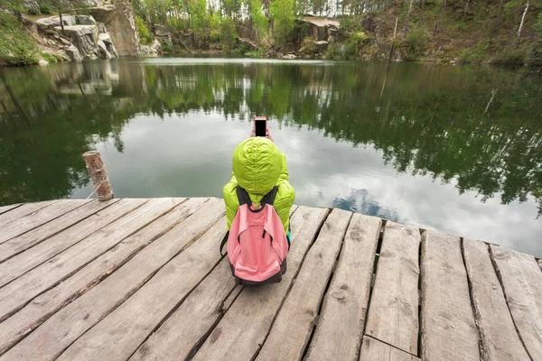 Uma viajante feminina está sentada em um cais de madeira perto de um belo lago em uma floresta de pinheiros e tirar fotos no celular . — Fotografia de Stock