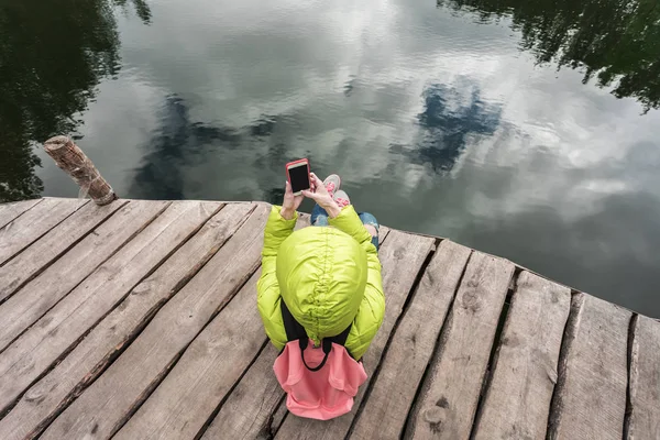 女性旅行者坐在附近一个美丽的湖，在一片松树林和拍照手机上的木码头上. — 图库照片