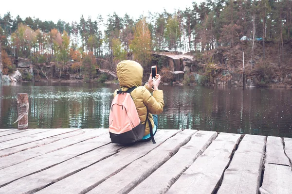 女性旅行者坐在附近一个美丽的湖，在一片松树林和拍照手机上的木码头上. — 图库照片