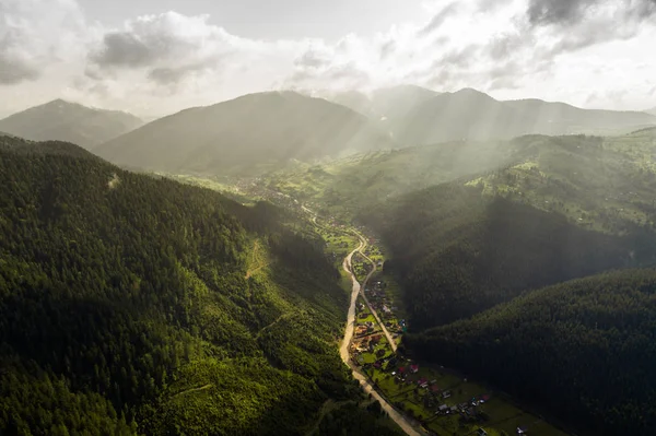 Nádherný panoramatický výhled ze vzduchu do údolí s vesnicí v Karpatských horách s mraky a sluneční paprsky v popředí — Stock fotografie