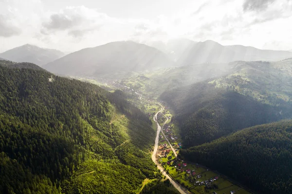 Nádherný panoramatický výhled ze vzduchu do údolí s vesnicí v Karpatských horách s mraky a sluneční paprsky v popředí — Stock fotografie
