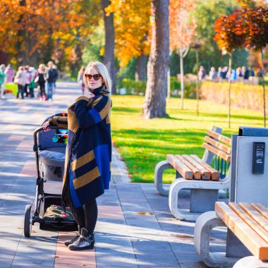 Genç bir anne bir bebek arabası ile sonbahar parkta yürüyor