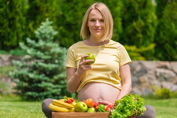 Çimenlerin üzerinde oturan ve bir elma elinde tutan hamile kadın — Stok fotoğraf