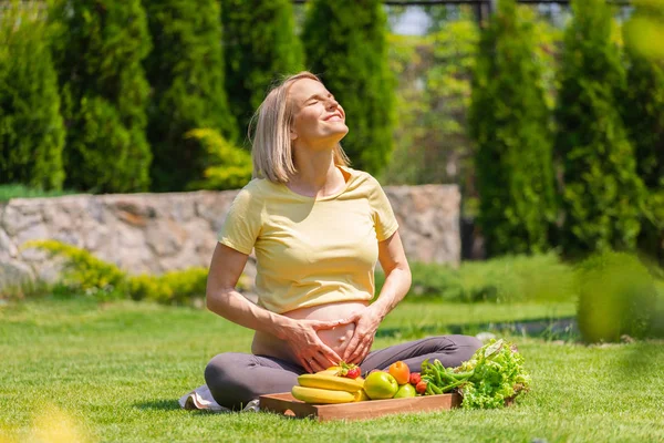 Una mujer embarazada se sienta en la hierba frente a una bandeja de frutas y retrata el corazón con sus manos sobre su vientre — Foto de Stock
