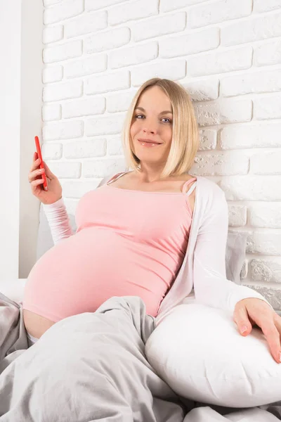 Felice donna incinta sorridente si trova a letto e guarda il telefono cellulare — Foto Stock