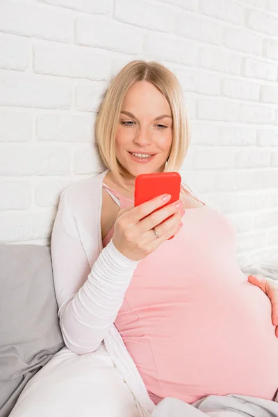 Feliz mujer embarazada sonriente se encuentra en la cama y mira el teléfono móvil —  Fotos de Stock