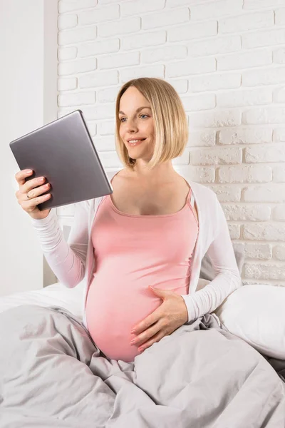 Szczęśliwy uśmiechający się kobieta w ciąży leży w łóżku i patrzy na tablecie. — Zdjęcie stockowe