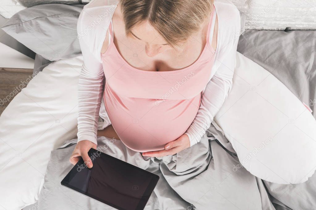 Happy smiling pregnant woman lies in bed and looks at tablet.