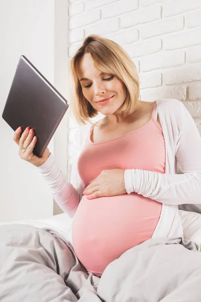 Glücklich lächelnde Schwangere liegt im Bett und blickt auf Tablet. — Stockfoto