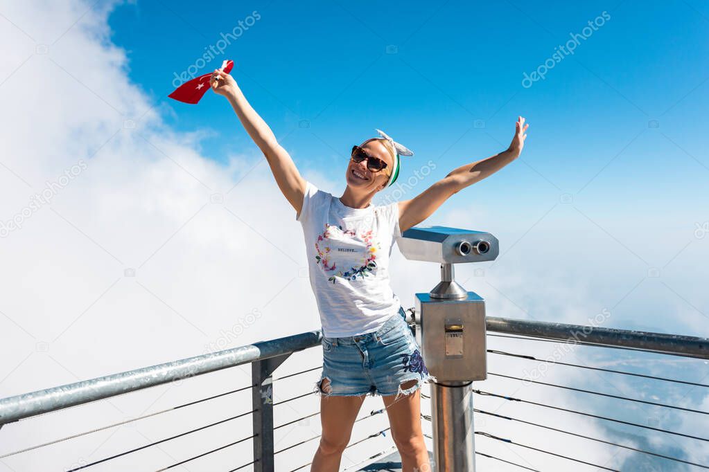 Young woman tourist stands on a viewing platform of Tahtali Mountain in Kemer with a red Turkey flag, Turkey