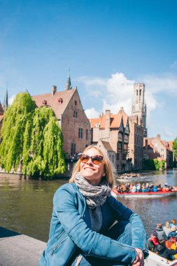 Belçika, Bruges 'de bir kanalı olan ünlü bir turist bölgesinin arka planında güzel bir genç kız oturuyor.