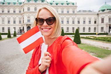Elinde Avusturya bayrağı olan genç ve mutlu bir bayan turist Avusturya, Viyana 'daki Belvedere Sarayı' nın arka planında selfie çekiyor.