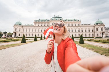 Elinde Avusturya bayrağı olan genç ve mutlu bir bayan turist Avusturya, Viyana 'daki Belvedere Sarayı' nın arka planında selfie çekiyor.