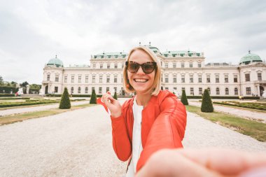 Elinde Avusturya bayrağı olan genç ve mutlu bir bayan turist Avusturya, Viyana 'daki Belvedere Sarayı' nın arka planında selfie çekiyor.