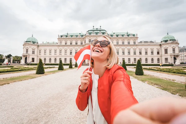 Uma jovem turista feliz com uma bandeira austríaca em suas mãos tira uma foto selfie no cenário do Palácio Belvedere em Viena, Áustria — Fotografia de Stock