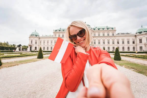 Una joven y feliz turista con una bandera austriaca en sus manos toma una foto selfie contra el telón de fondo del Palacio Belvedere en Viena, Austria — Foto de Stock