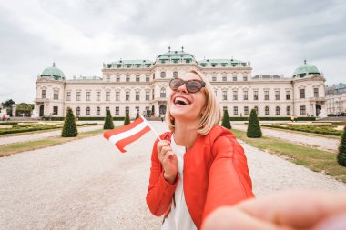 Elinde Avusturya bayrağı olan genç ve mutlu bir bayan turist Avusturya, Viyana 'daki Belvedere Sarayı' nın arka planında selfie çekiyor.