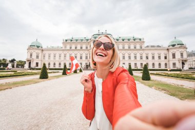 Elinde Avusturya bayrağı olan genç ve mutlu bir bayan turist Avusturya, Viyana 'daki Belvedere Sarayı' nın arka planında selfie çekiyor.