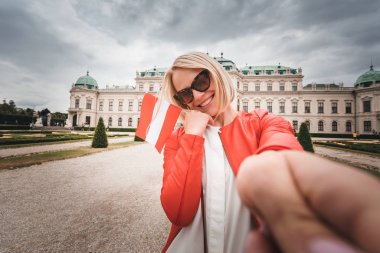 Elinde Avusturya bayrağı olan genç ve mutlu bir bayan turist Avusturya, Viyana 'daki Belvedere Sarayı' nın arka planında selfie çekiyor.