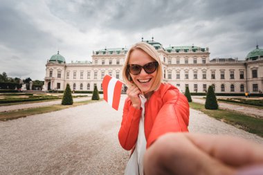 Elinde Avusturya bayrağı olan genç ve mutlu bir bayan turist Avusturya, Viyana 'daki Belvedere Sarayı' nın arka planında selfie çekiyor.