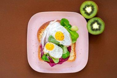 Sağlıklı kahvaltı sandviçi konsepti. Kızarmış yumurtalı ekmek ve kahverengi arka planda pembe tabakta yeşil salata. Beslenme yemeği.