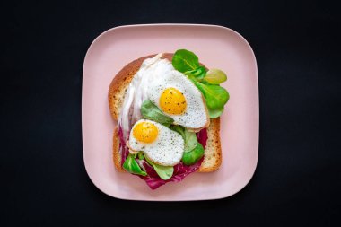 Sağlıklı kahvaltı sandviçi konsepti. Kızarmış yumurtalı ekmek ve siyah arka planda pembe tabakta yeşil salata. Beslenme yemeği.