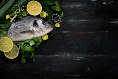 Siyah ahşap zemin üzerinde limon ve bitkilerle taze ve çiğ dorada balığı, üst görünüm ve boşluk kopyala