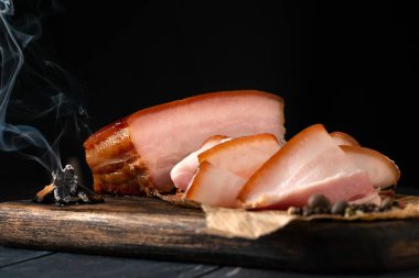 Tütsülenmiş domuz pastırması, bir parça duman ve ahşap bir tahtada dilimlenmiş.