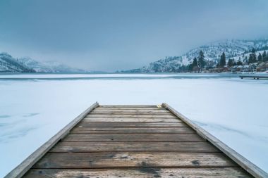 Winter landscape of wooden dock in foreground on frozen lake with snow covered mountains and fog in background clipart