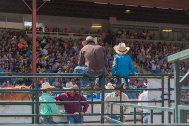 Williams Lake, British Columbia/Canada - July 2, 2016: man and boy sit on fence with a group of cowboys, watching the action at the 90th Williams Lake Stampede, an internationally famous event. clipart