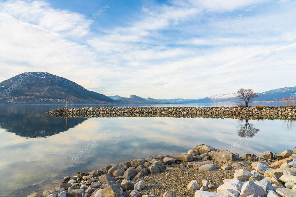 Breakwater, mountains, and sky reflected in water of calm lake with rocky shoreline