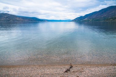 Çakıl taşı sahili, sakin göl, dağlar ve Antler 'in Sahili, Peachland, British Columbia, Kanada' da bulutlu gökyüzü