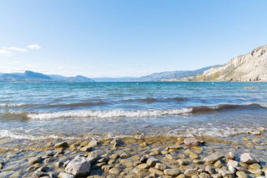 Dalgalar Okanagan Gölü 'nün kayalık kıyı şeridine çarpıyor. Mavi gökyüzü ve uzak sıradağlar görünüyor.