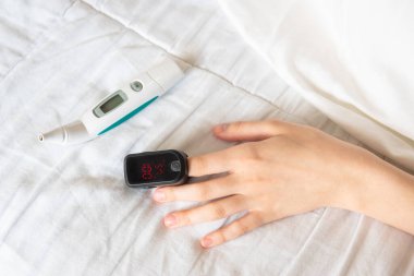 Yatağın üzerinde dijital termometreyle parmak üstüne nabız oksimetresi takan bir kadın eli.
