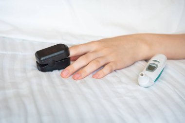 Kadının eli dijital termometreyle yatakta dinleniyor ve parmağında nabız oksimetresi var.