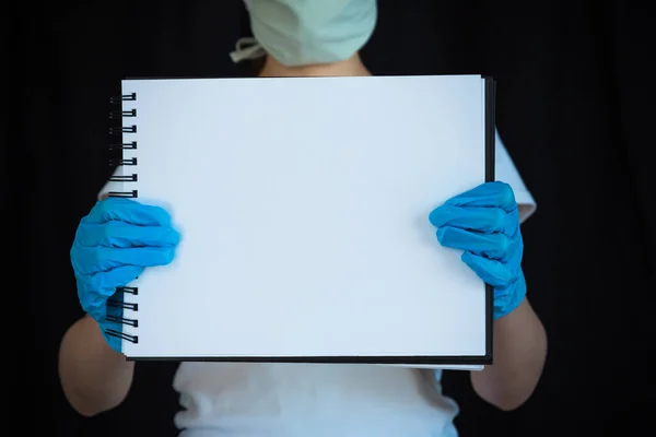 Woman wearing face mask and surgical gloves, holding blank note pad with black background