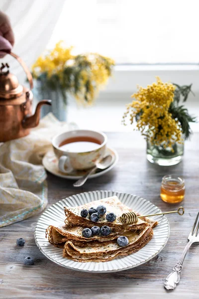 Ein Teller mit Pfannkuchen mit Blaubeeren auf einem Holztisch. Im Hintergrund eine Tasse Tee und ein Strauß Frühlingsblumen am Fenster. — Stockfoto