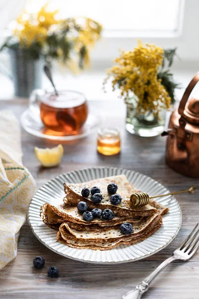 Ein Teller mit Pfannkuchen mit Blaubeeren auf einem Holztisch. Im Hintergrund eine Tasse Tee und ein Strauß Frühlingsblumen am Fenster. — Stockfoto
