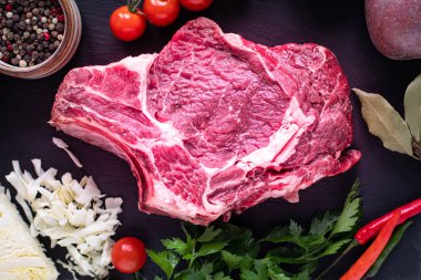 Çiğ sulu biftek, siyah tahtada ızgara yapmaya hazır. Kemikte biftek, ahşap bir kesim tahtasının üzerinde vişneli domates, acı biber ve bitkilerle birlikte dana eti. en üstteki görünüm