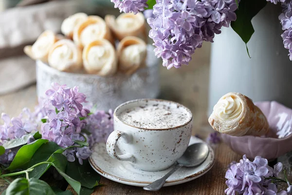 Cup of cappuccino and cake cones from puff pastry with vanilla cream in a metal box in spring still life with a bouquet of lilacs on a wooden table