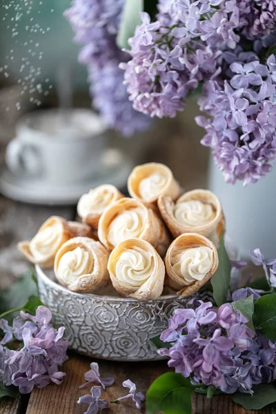 Baharda metal bir kutuda vanilya kremalı poğaça boynuzları ve ahşap bir masada bir buket leylak.