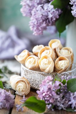 Baharda metal bir kutuda vanilya kremalı poğaça boynuzları ve ahşap bir masada bir buket leylak.