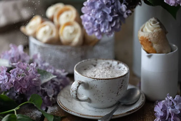 Cup of cappuccino and cake cones from puff pastry with vanilla cream in a metal box in spring still life with a bouquet of lilacs on a wooden table