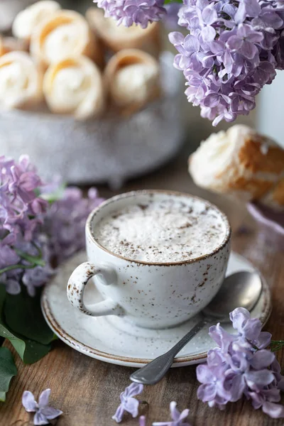 Cup of cappuccino and cake cones from puff pastry with vanilla cream in a metal box in spring still life with a bouquet of lilacs on a wooden table