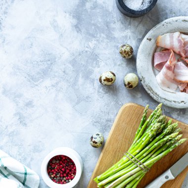 Gri beton arka planda yemek pişirmek için gerekli malzemeler. Bir demet taze kuşkonmaz, yumurta ve pastırma. Üst Manzara. Uzayı kopyala Kare