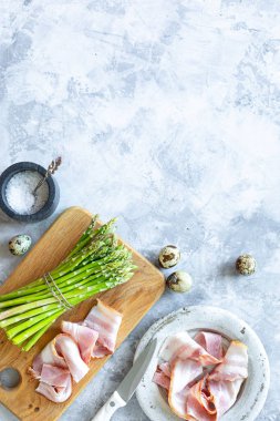 Gri beton arka planda yemek pişirmek için gerekli malzemeler. Bir demet taze kuşkonmaz, yumurta ve pastırma. Üst Manzara. Uzayı kopyala Dikey