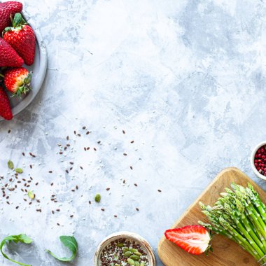 Gri beton arka planda yemek pişirmek için gerekli malzemeler. Bir demet taze kuşkonmaz, çilek, roka. Üst Manzara. Uzayı kopyala Kare